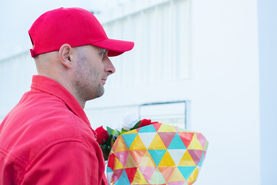 A delivery man wearing a red cap and matching red jacket is holding a gift box wrapped in brightly coloured paper with a geometric triangle pattern in shades of yellow, blue, red, and green. The man i