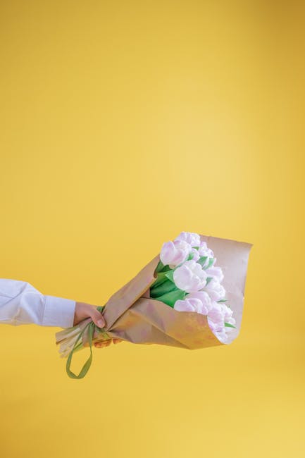 A hand holding a bouquet of soft pink tulips wrapped in brown craft paper with green accents, presented against a bright yellow background. The tulips have smooth, rounded petals and fresh green stems