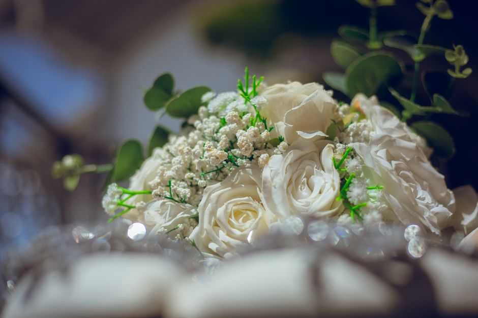 A close-up view of a floral arrangement featuring white roses, accented with small white baby's breath flowers and lush green eucalyptus leaves. The bouquet is arranged with the roses as the focal poi