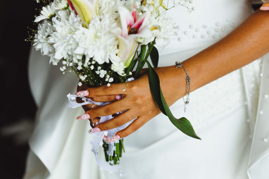 Close-up of a bride's hand holding a floral bouquet consisting of white lilies, small white chrysanthemums, and baby's breath, wrapped with a white ribbon. The bride's hand displays manicured nails wi