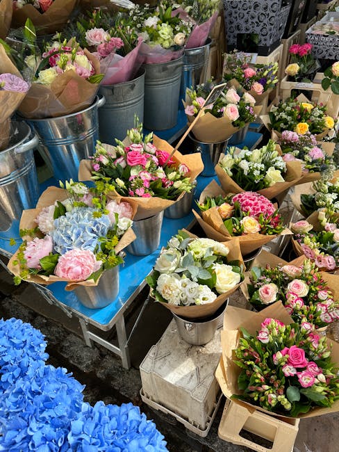 An array of fresh floral arrangements displayed on a blue table at Northolt Road Market, featuring pink, white, and blue flowers arranged in small wrapped bouquets and metal buckets. The bouquets incl