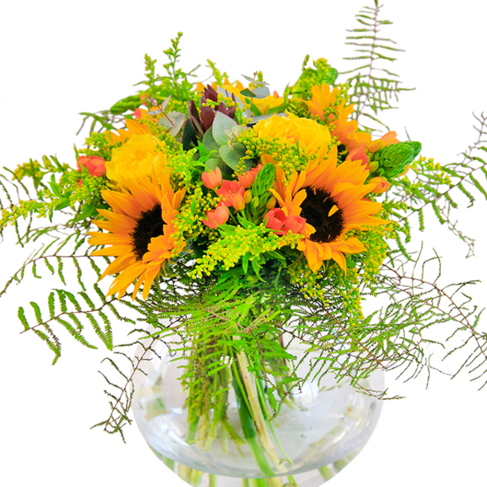 Pure sunshine radiates from this handcrafted Greenford bouquet-a joyful celebration of golden hues, abundant textures, and a welcoming West London spirit. The eye is instantly drawn to radiant sunflowers, their deep chocolate centres framed by generous yellow petals that appear to glow with afternoon light. Clustered around them, golden-yellow garden roses open gracefully in tightly whorled spirals, releasing a delicate, sweet fragrance reminiscent of nearby Brent Valley's summer gardens. Here and there, shiny red Hypericum berries pop from the composition, their vivid hue drawing out the vibrancy of the surrounding blooms. Feathery fronds of fern cascade over the edge of a pristine, globe-shaped glass vase, their gentle lines softening the overall design and inviting the touch-like foliage brushed by a passing breeze on Greenford Broadway. Sprigs of light green filler, tinged with the tiniest yellow blossoms-perhaps goldenrod-add further dimension, while the bluish-green sheen of eucalyptus leaves lends a modern, natural accent. The clear vase magnifies the beauty below: tangled green stems and shimmering water, a clean and vital foundation for this exuberant display. Every element of the bouquet, from bold sunflower to delicate greenery, has been selected to bring moments of radiant joy into Greenford homes-whether marking a milestone or simply brightening the day of someone special.